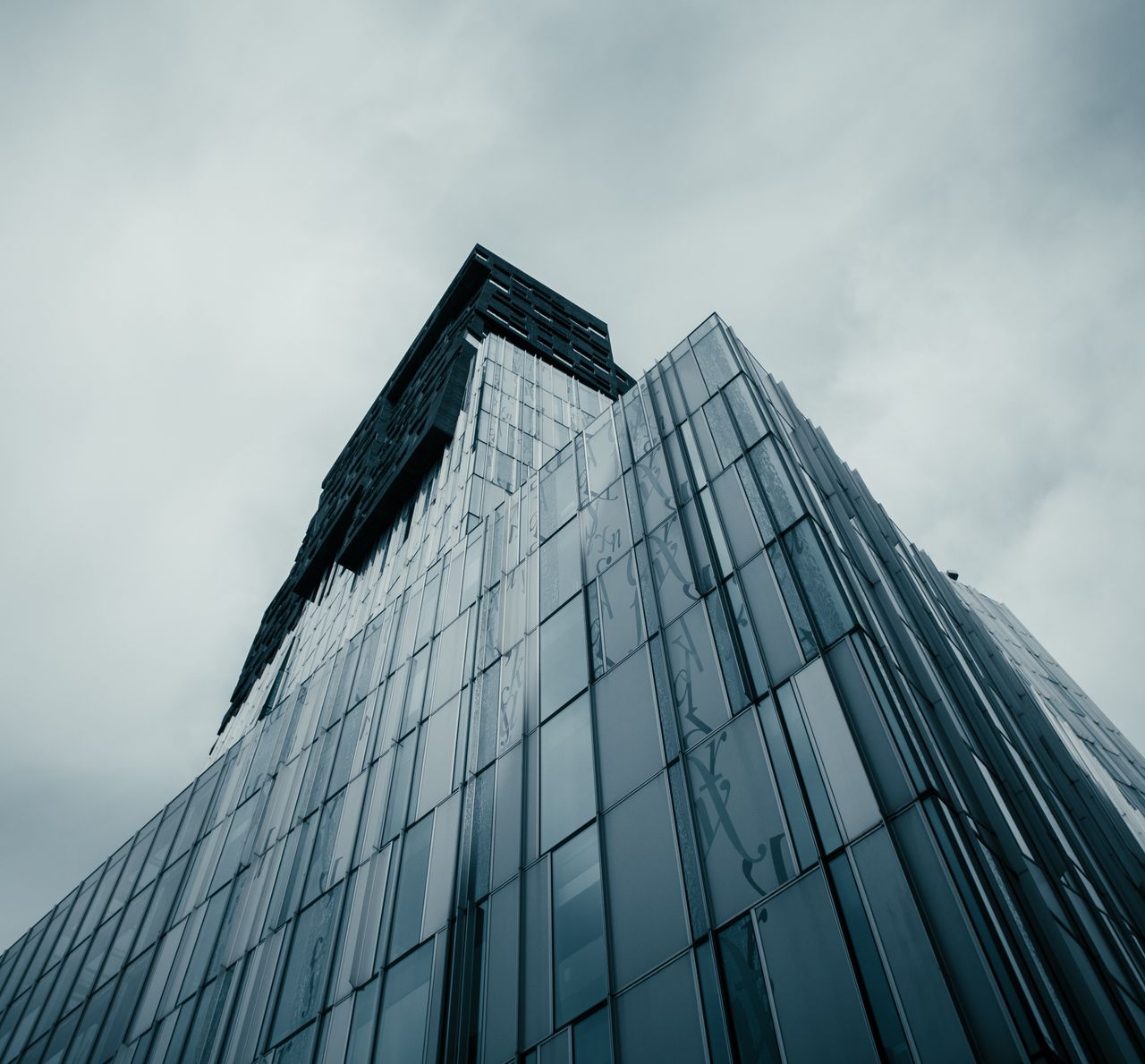 services-01 Low angle view of a modern glass skyscraper under a cloudy sky, showcasing contemporary architecture.